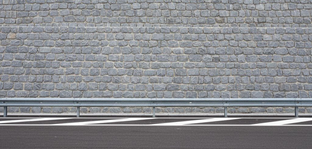 Muro di contenimento in pietra grigia a vista, di nuova costruzione, con guardrail metallico e asfalto in primo piano.