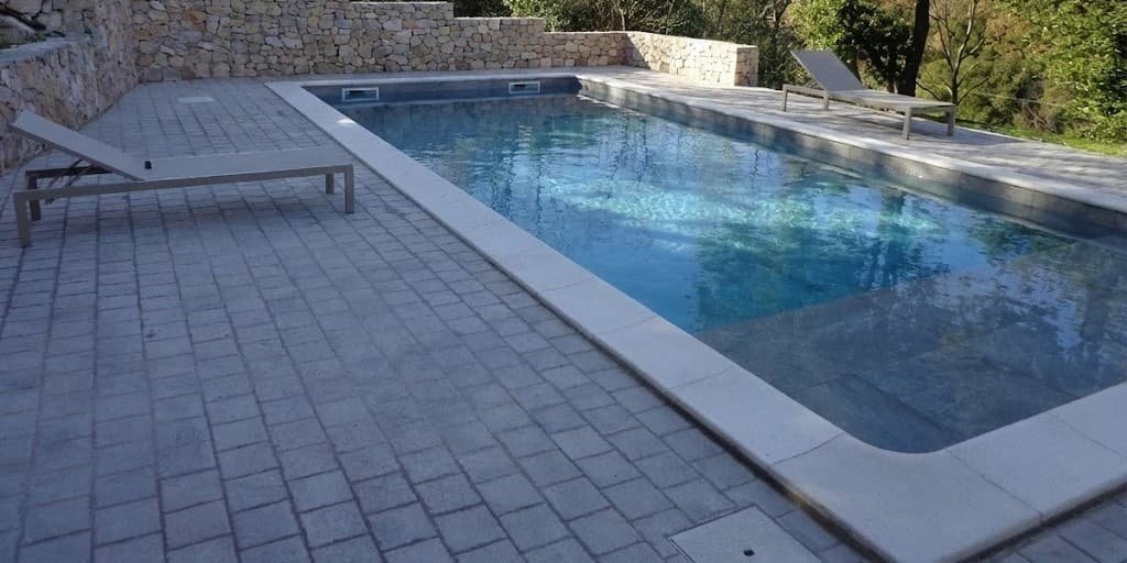 Piscina interrata rettangolare moderna con bordo perimetrale chiaro e solarium in pietra naturale, circondata da muretti di contenimento a secco in un contesto paesaggistico ligure.