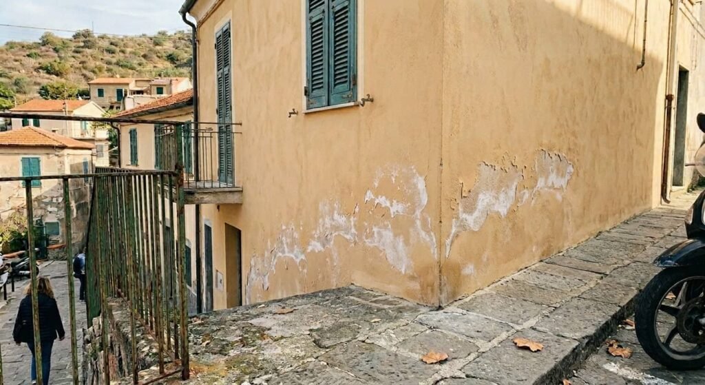 Distacco intonaco cristallizzazione sali mare liguria Prospetto di un edificio costiero che mostra un degrado murario avanzato: una banda orizzontale di efflorescenze bianche e intonaco scrostato rivela gli effetti della pressione di cristallizzazione dei sali marini. La muratura ammalorata evidenzia come l'evaporazione forzata dell'umidità salina spacchi i rivestimenti tradizionali, rendendo necessario un risanamento con sistemi macroporosi